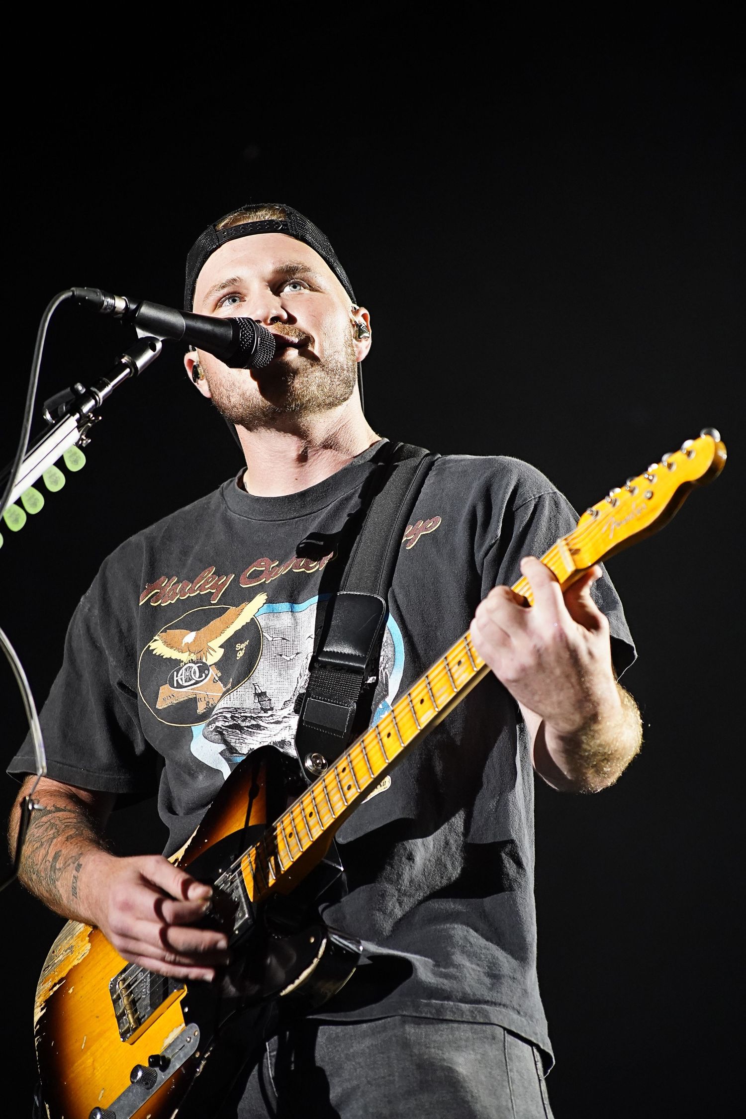 Musician performs with yellow electric guitar under stage lights wearing baseball cap.