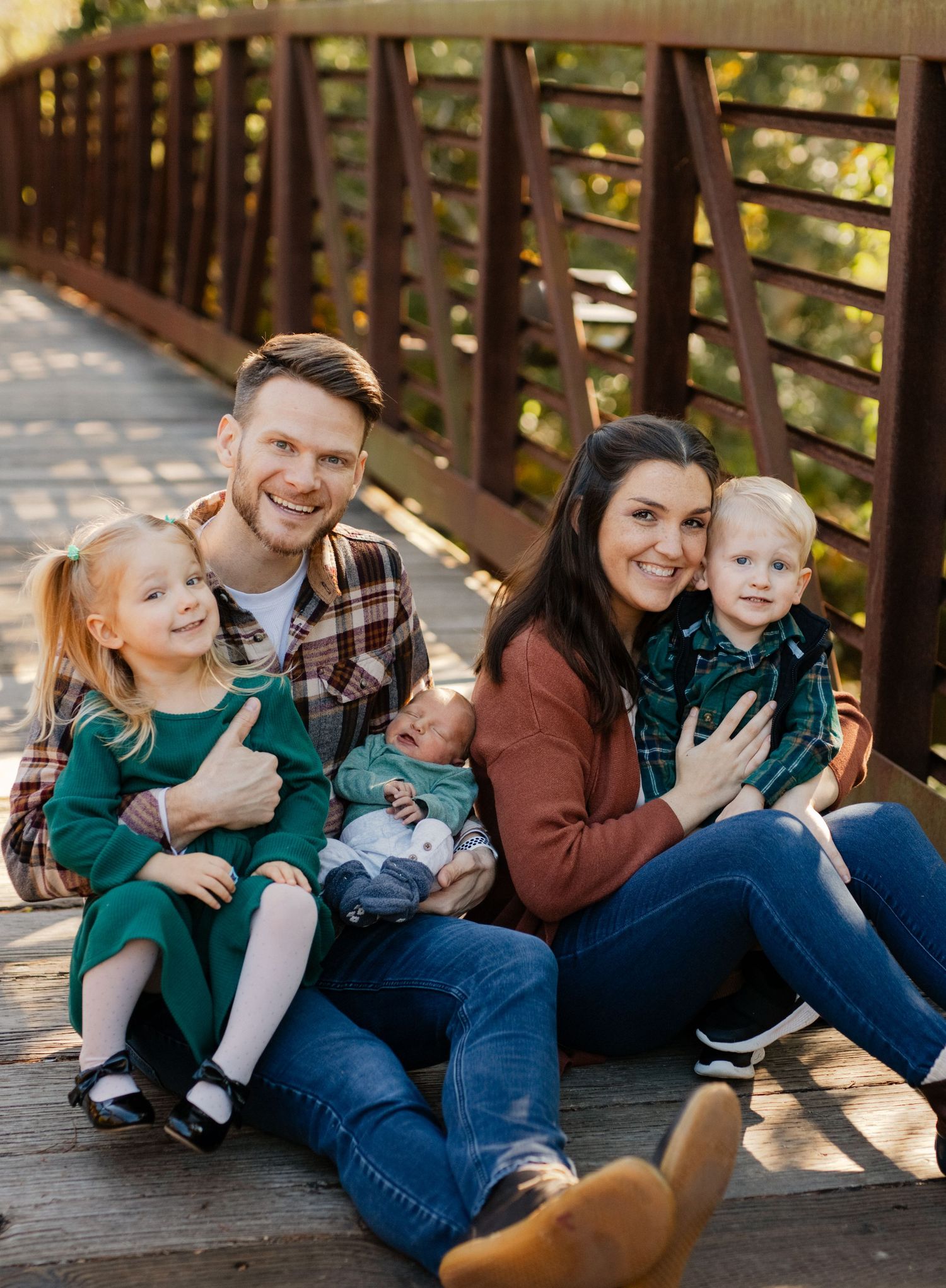 Family of four sits together on a wooden bridge for a casual outdoor portrait.