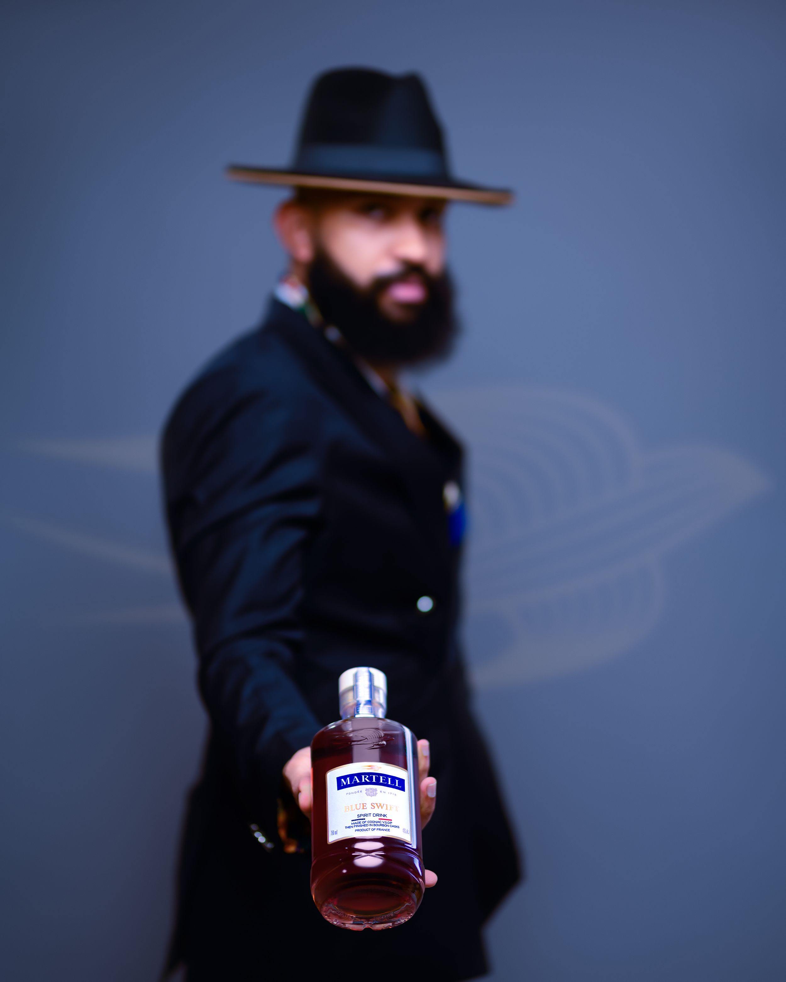A moody portrait of someone in a fedora holding a bottle against a blue background.