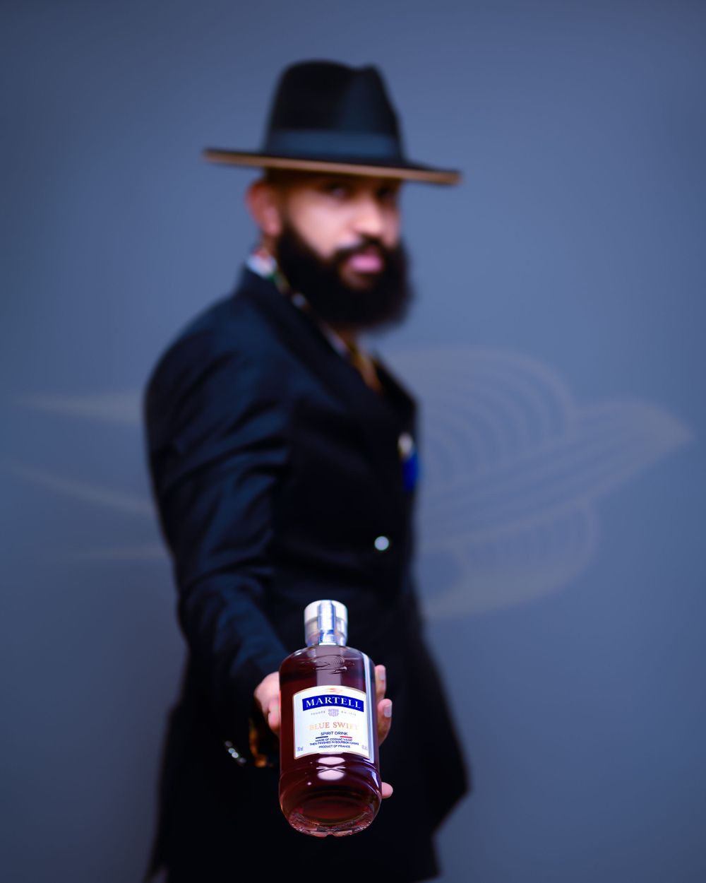 A moody portrait of someone in a fedora holding a bottle against a blue background.