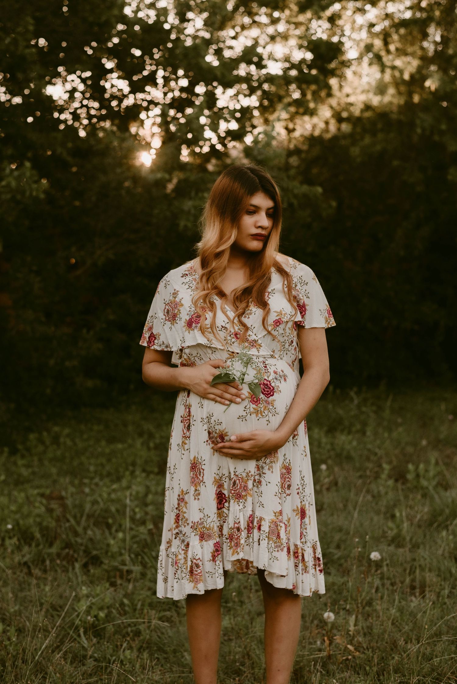 A pregnant person in a floral dress stands in a grassy field at sunset with sunlight filtering through trees.