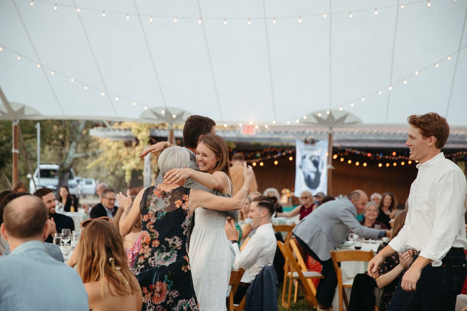 Back view of guests hugging during outdoor wedding reception under warm lighting.
