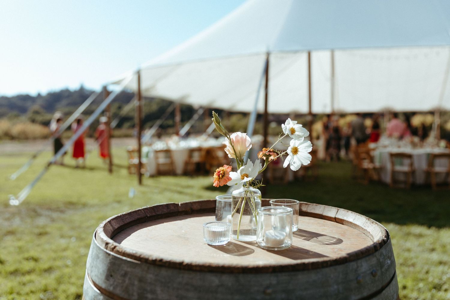 Mason jar with delicate flowers displayed on a wooden barrel under a white reception tent.