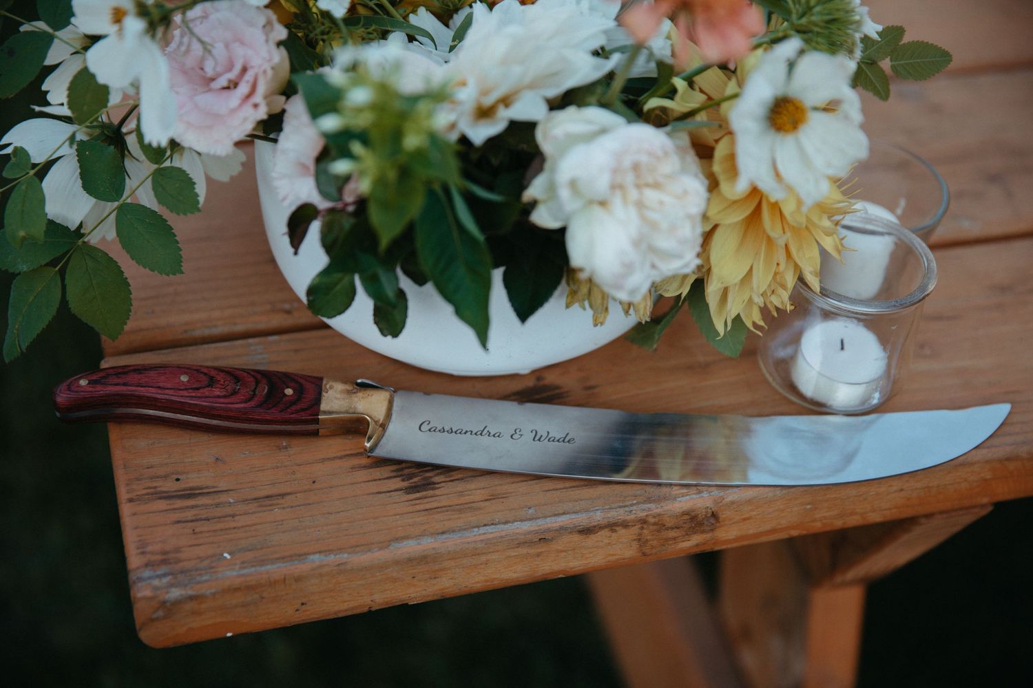 Wedding knife resting on wooden table next to floral centerpiece with roses and dahlias.