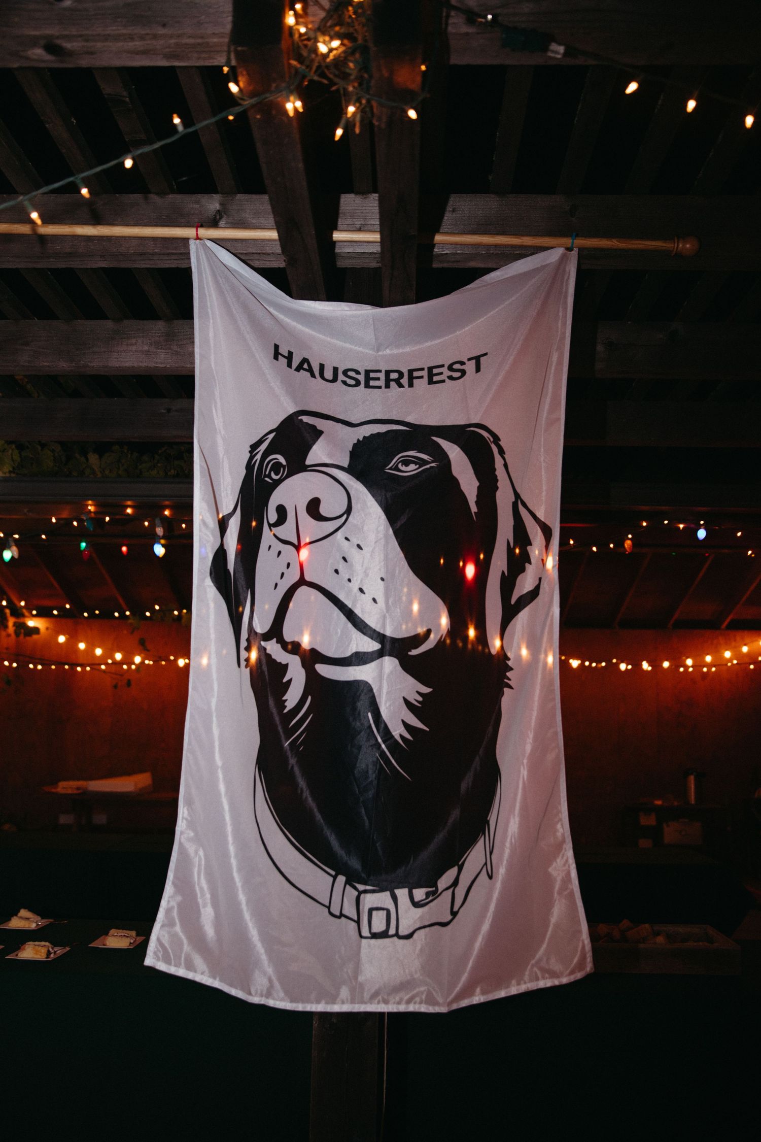 White banner with black and white dog illustration hanging from wooden beams with string lights in background.