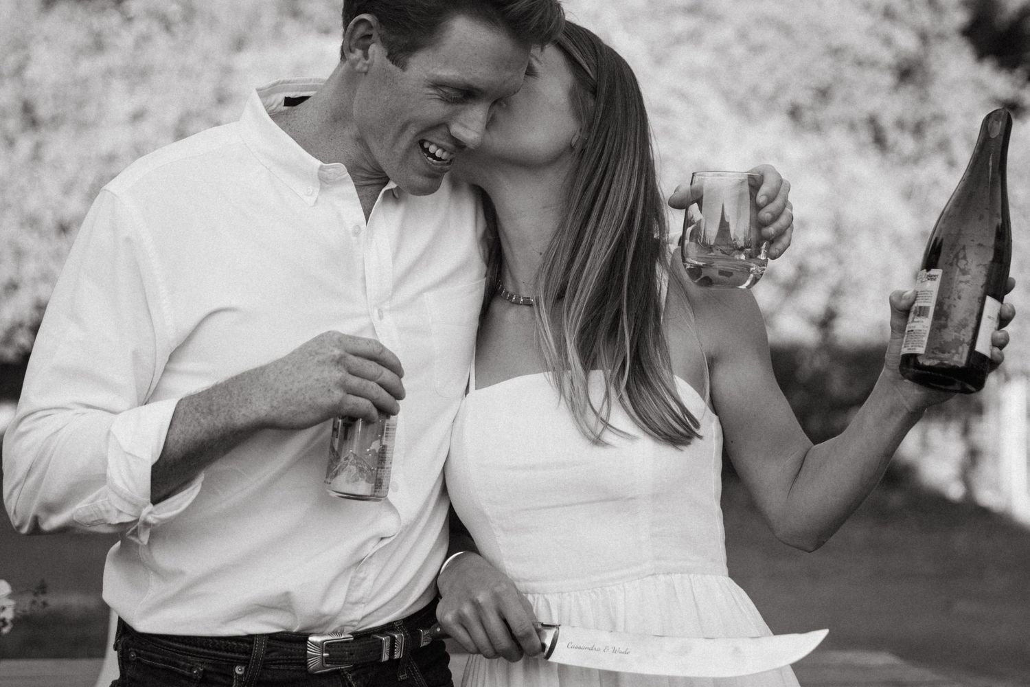 A black and white candid shot of two people sharing an intimate moment while holding drinks outdoors.