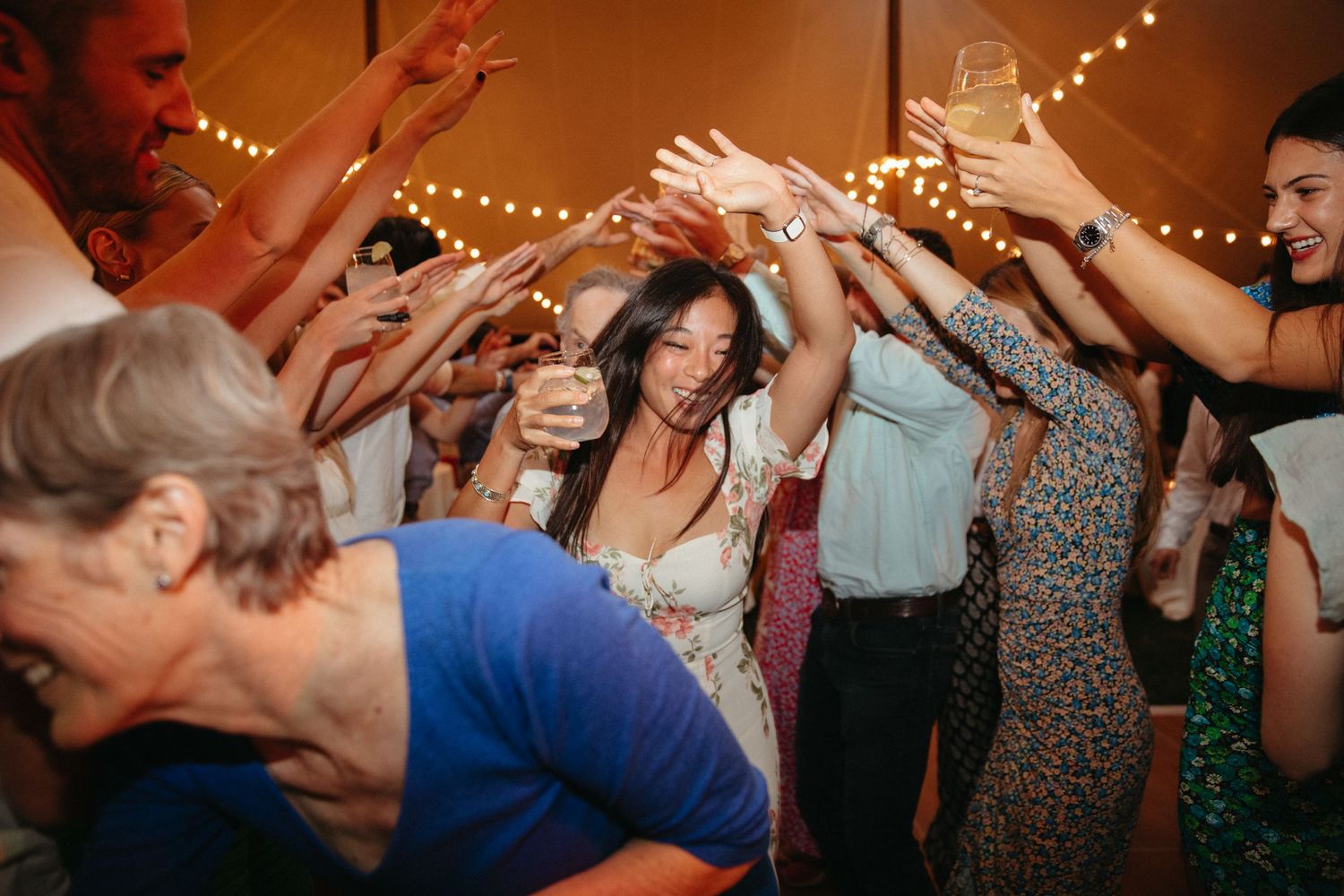 Energetic crowd dances with raised arms under string lights at wedding reception.