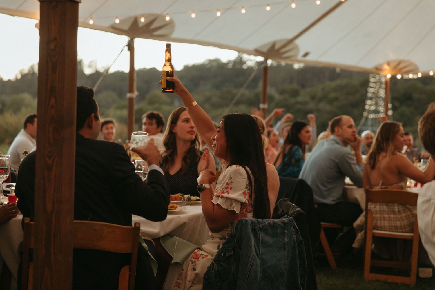 Wedding guests raise glasses in celebration under a tent with string lights at sunset.