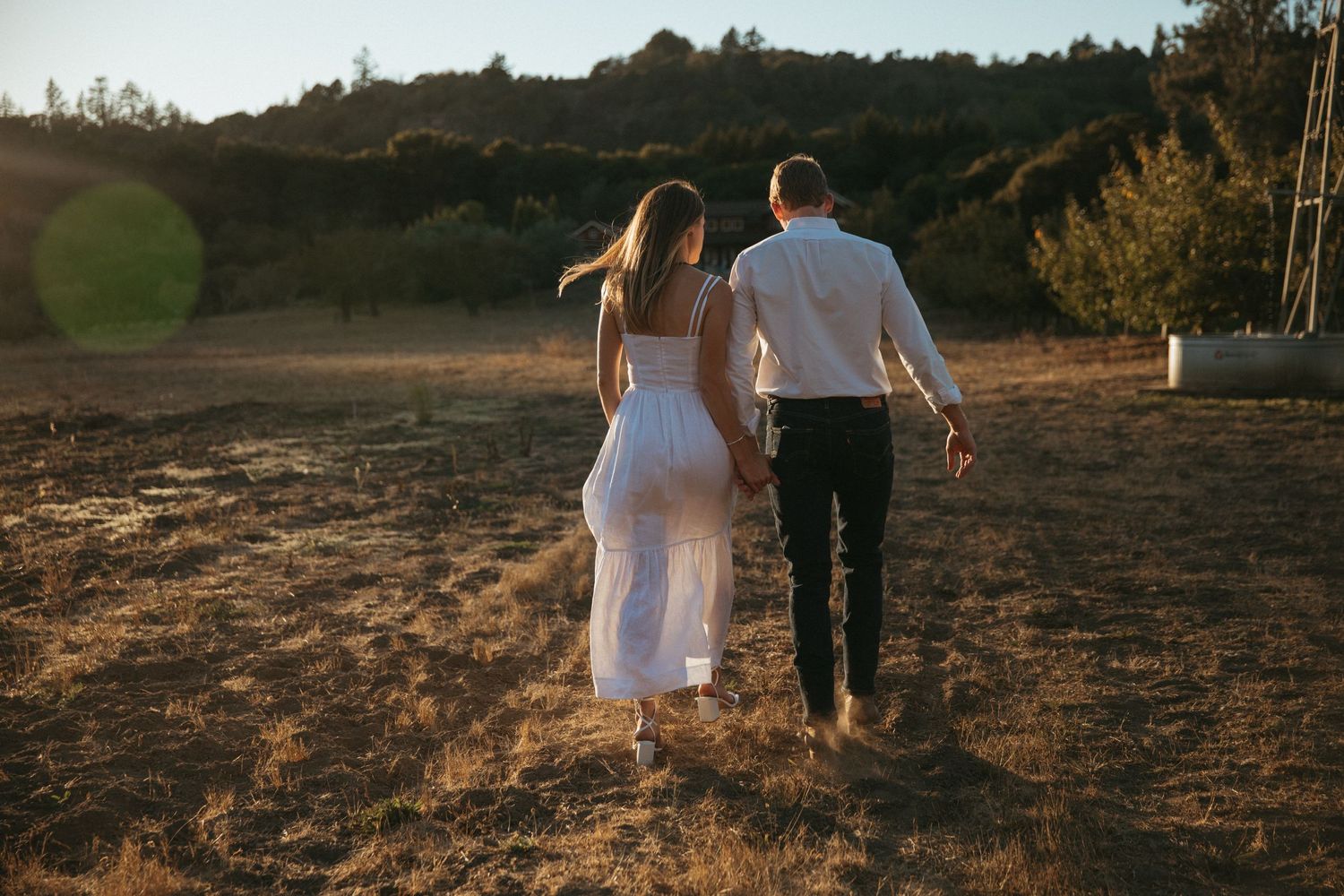 Couple in white and dark attire walking through golden sunlit field.