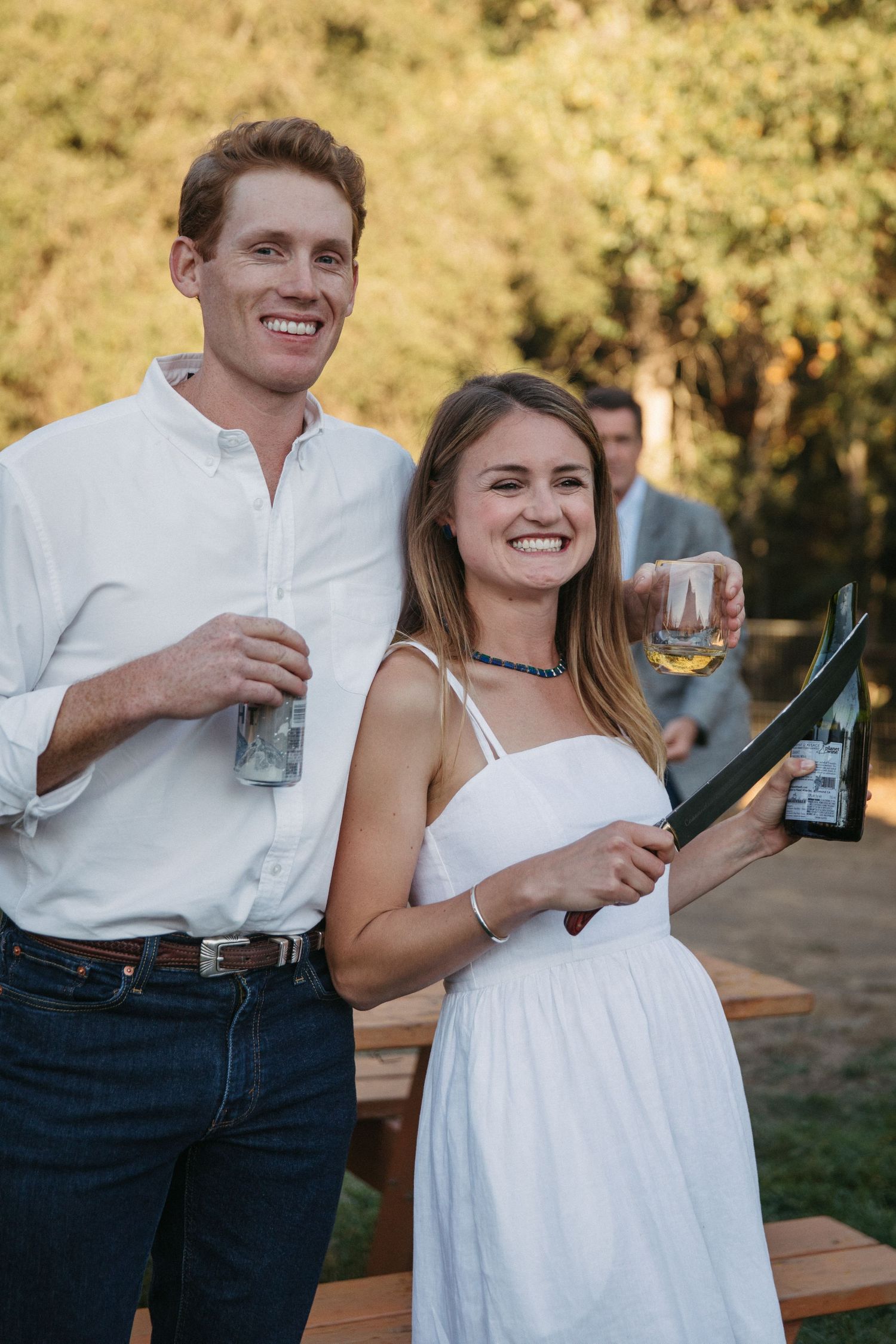 A couple poses with drinks at an outdoor celebration against a backdrop of golden trees.