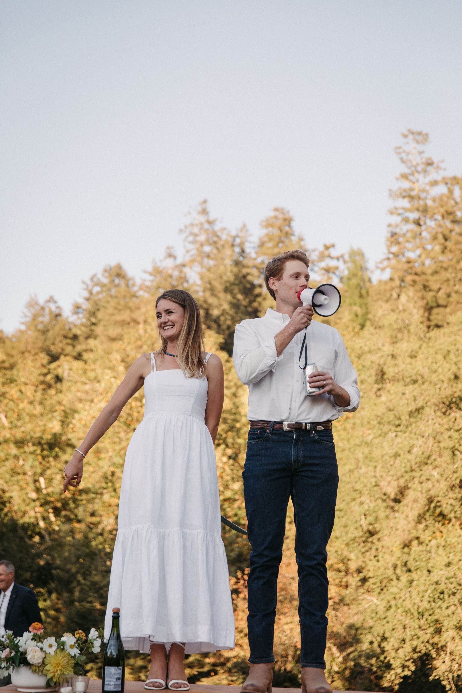 A couple stands on a platform addressing guests at an outdoor celebration with autumn trees in the background.