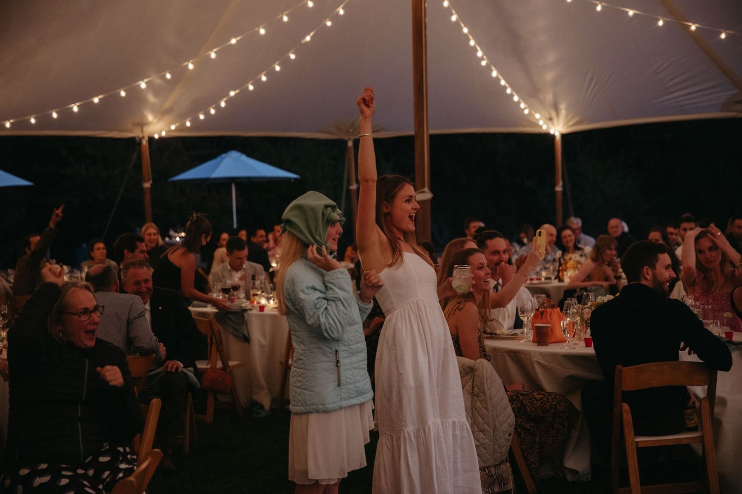 Wedding guests dance and cheer under twinkling lights at evening reception.