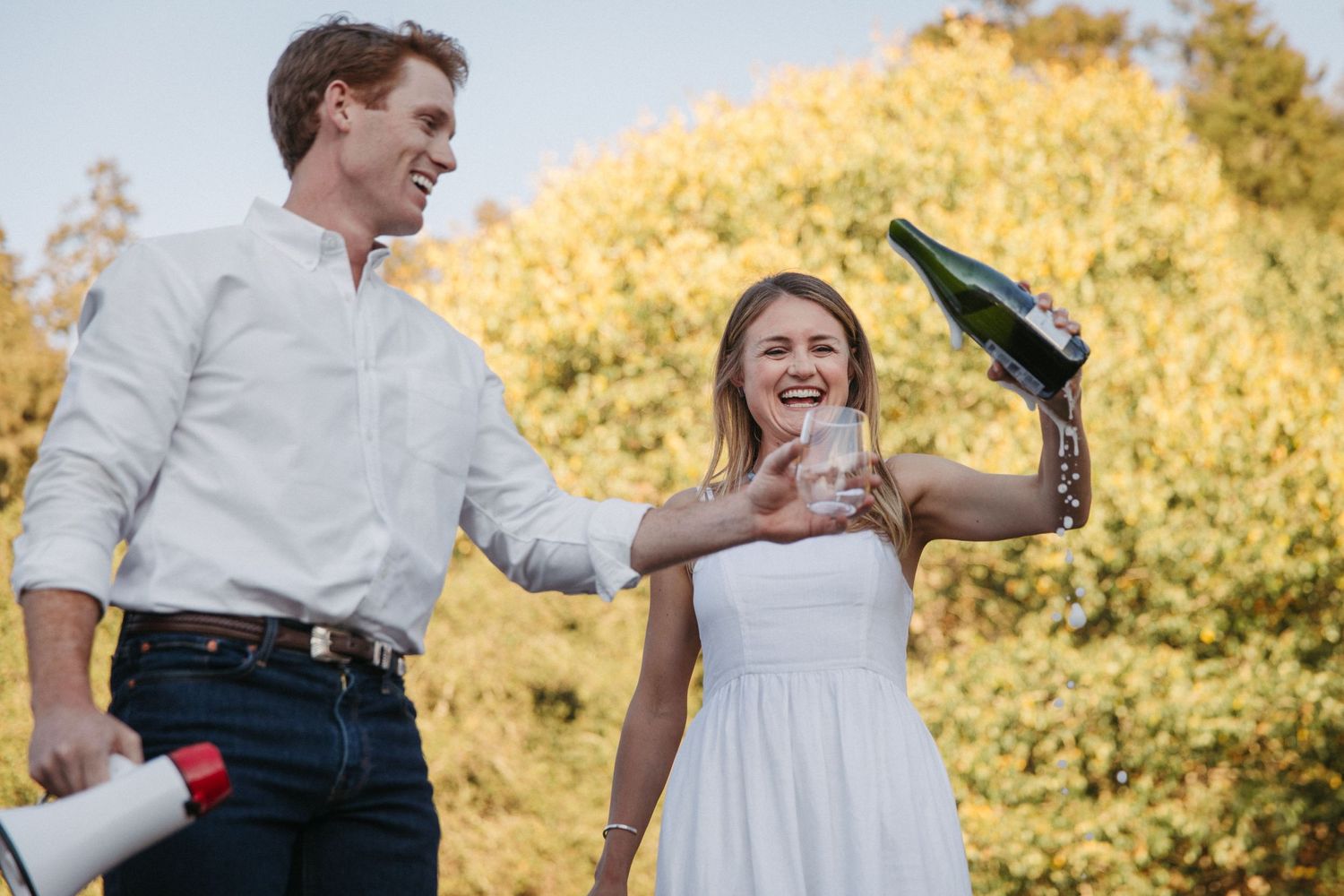 Two people laughing while performing a champagne celebration outdoors with autumn foliage.