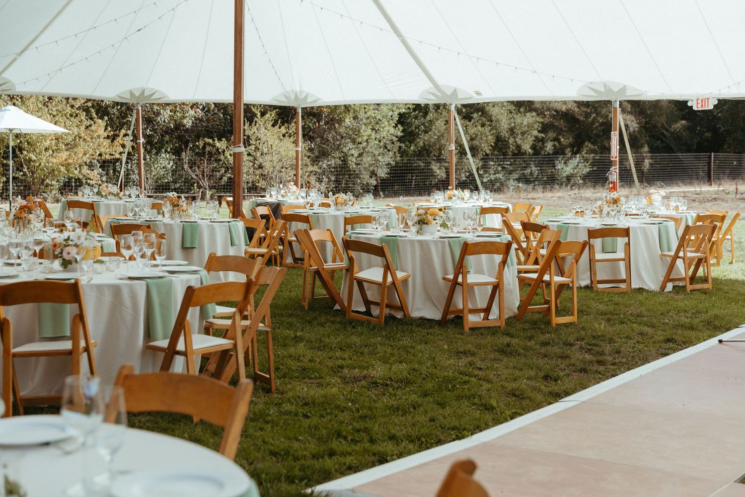 Outdoor wedding reception setup with white tables and wooden chairs under a tent.