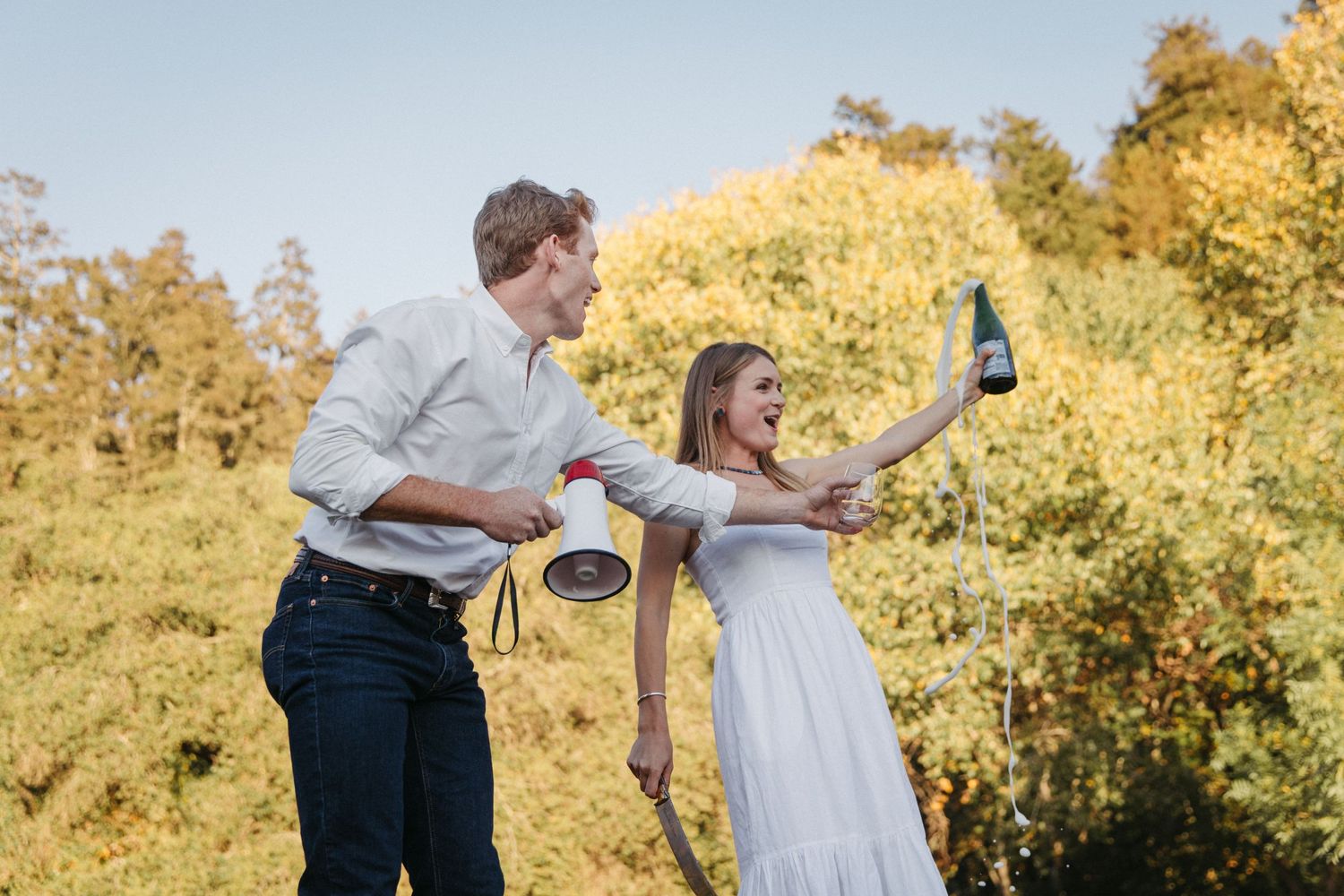 A couple poses playfully with drinks and a megaphone against a backdrop of golden autumn trees.