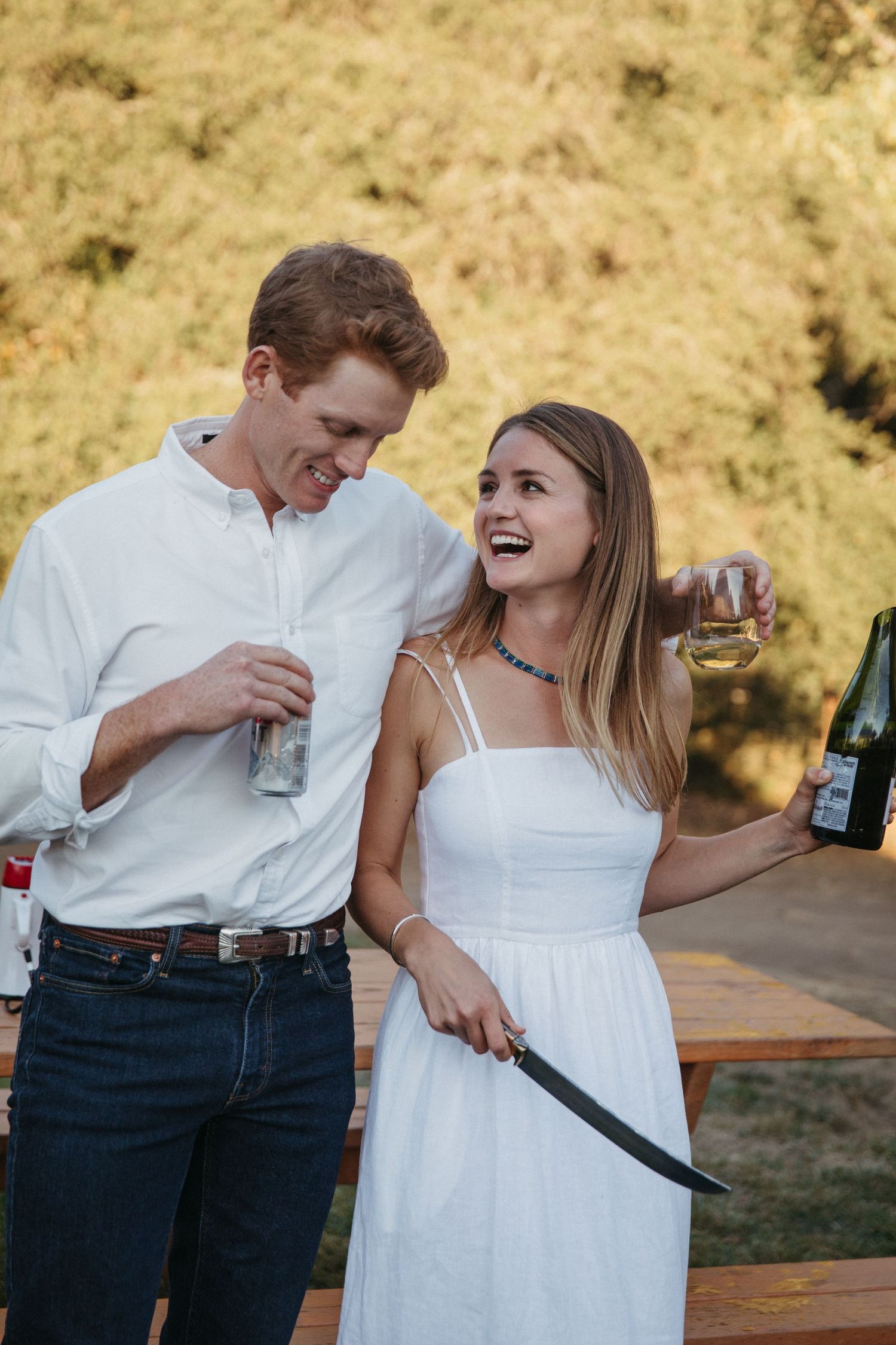A candid moment of two people laughing while holding drinks at an outdoor celebration.