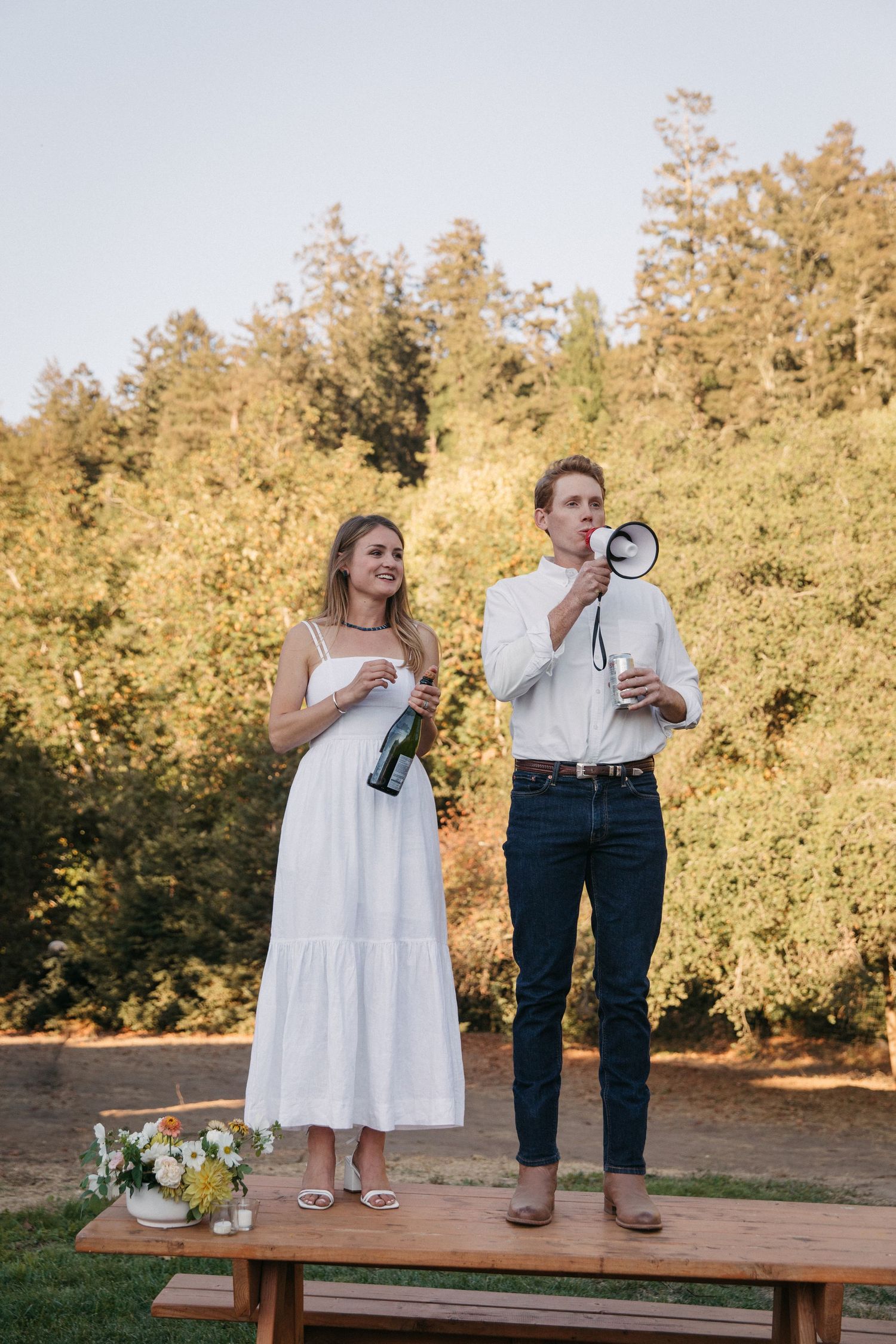 A couple makes speeches while standing on a wooden table at an outdoor event.