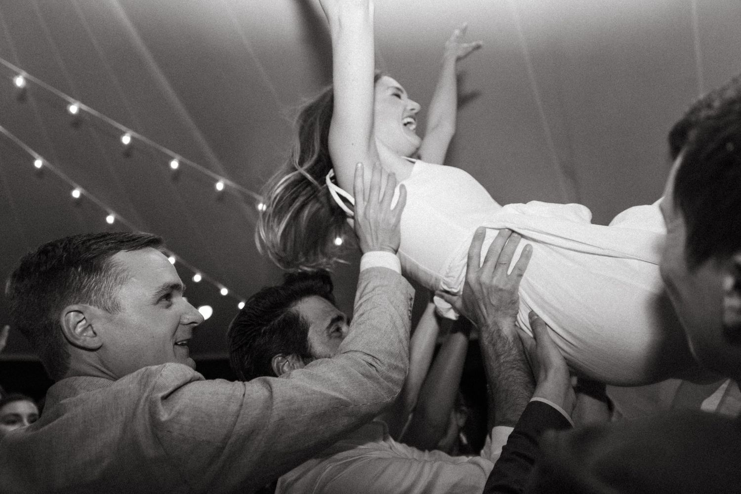 Wedding guests lift and celebrate during a lively dance party under string lights.