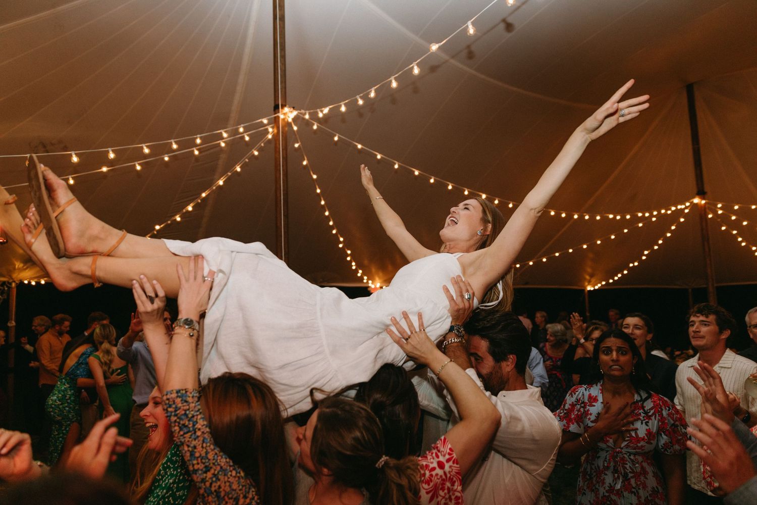 Wedding guests lift and support woman in white dress during reception dance party.