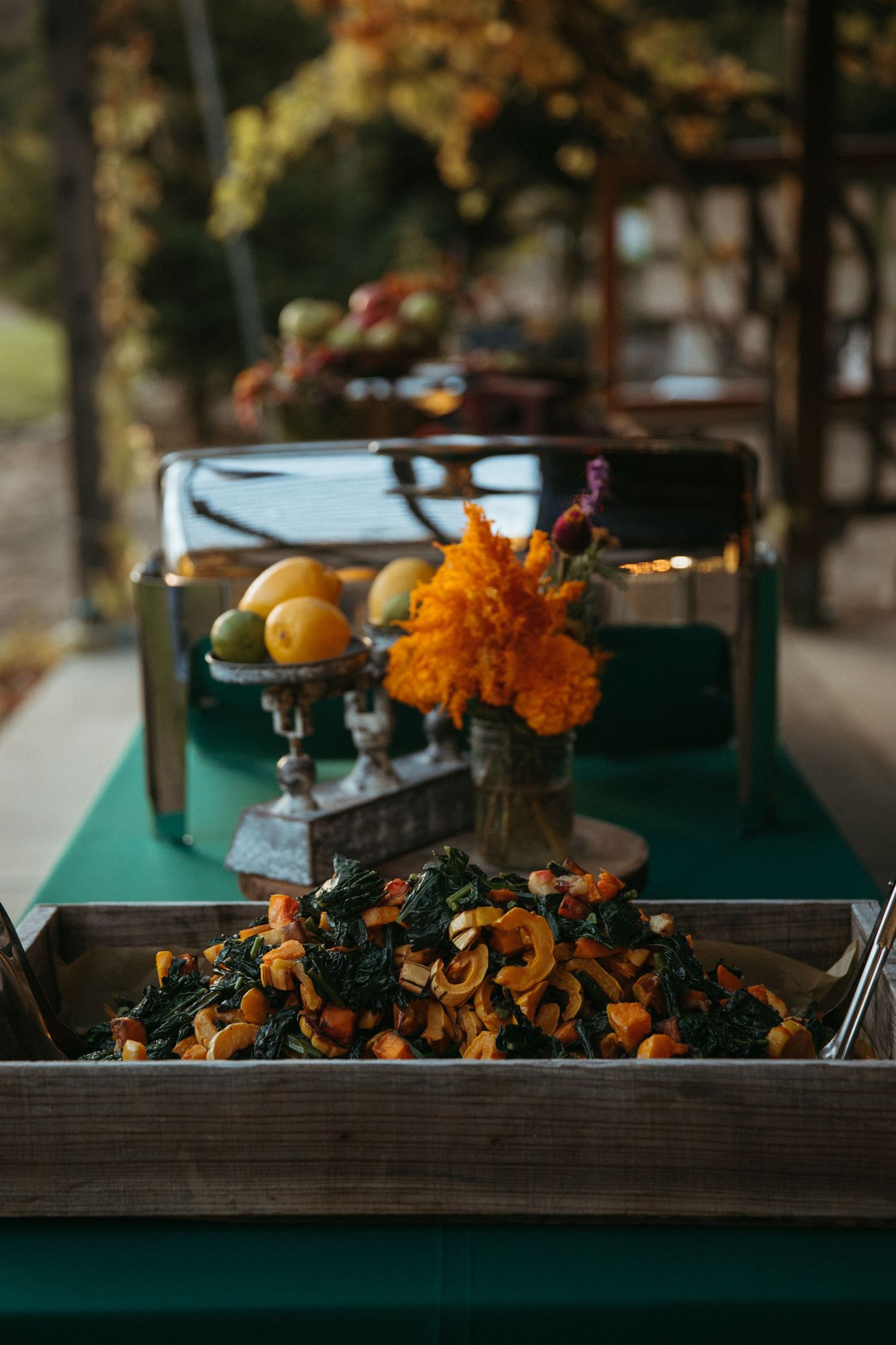 Rustic food display with orange flowers and dark greens on emerald green tablecloth.