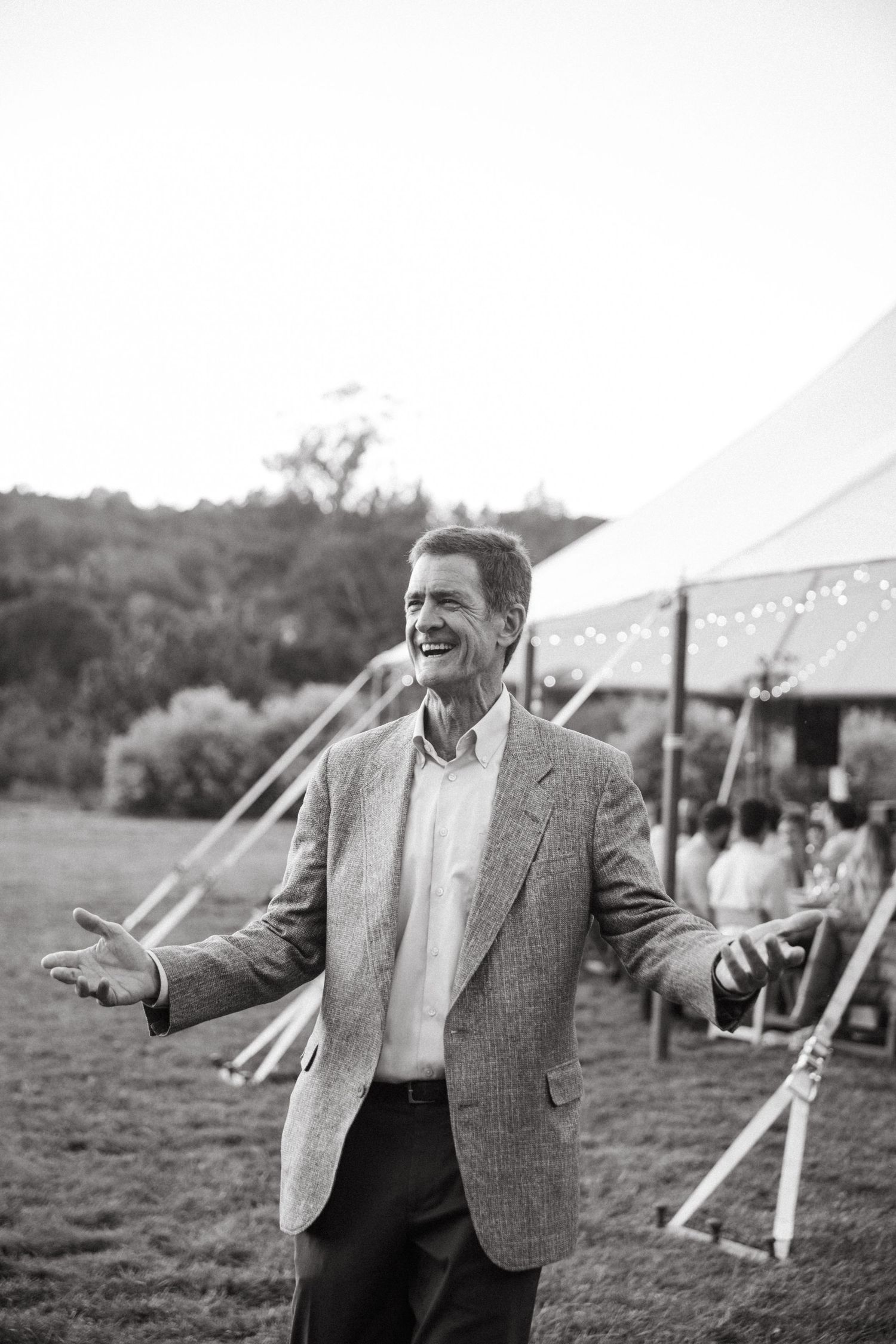 Wedding guest speaks joyfully outdoors with arms outstretched under white tent at outdoor reception.
