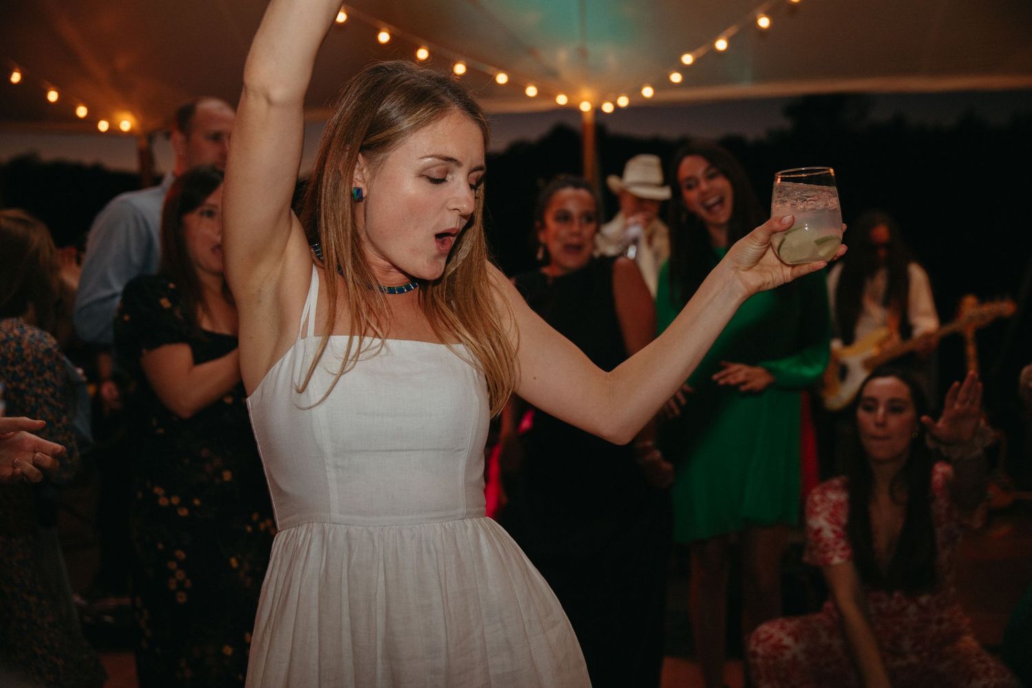 A guest in white dress dances energetically at wedding reception under string lights.