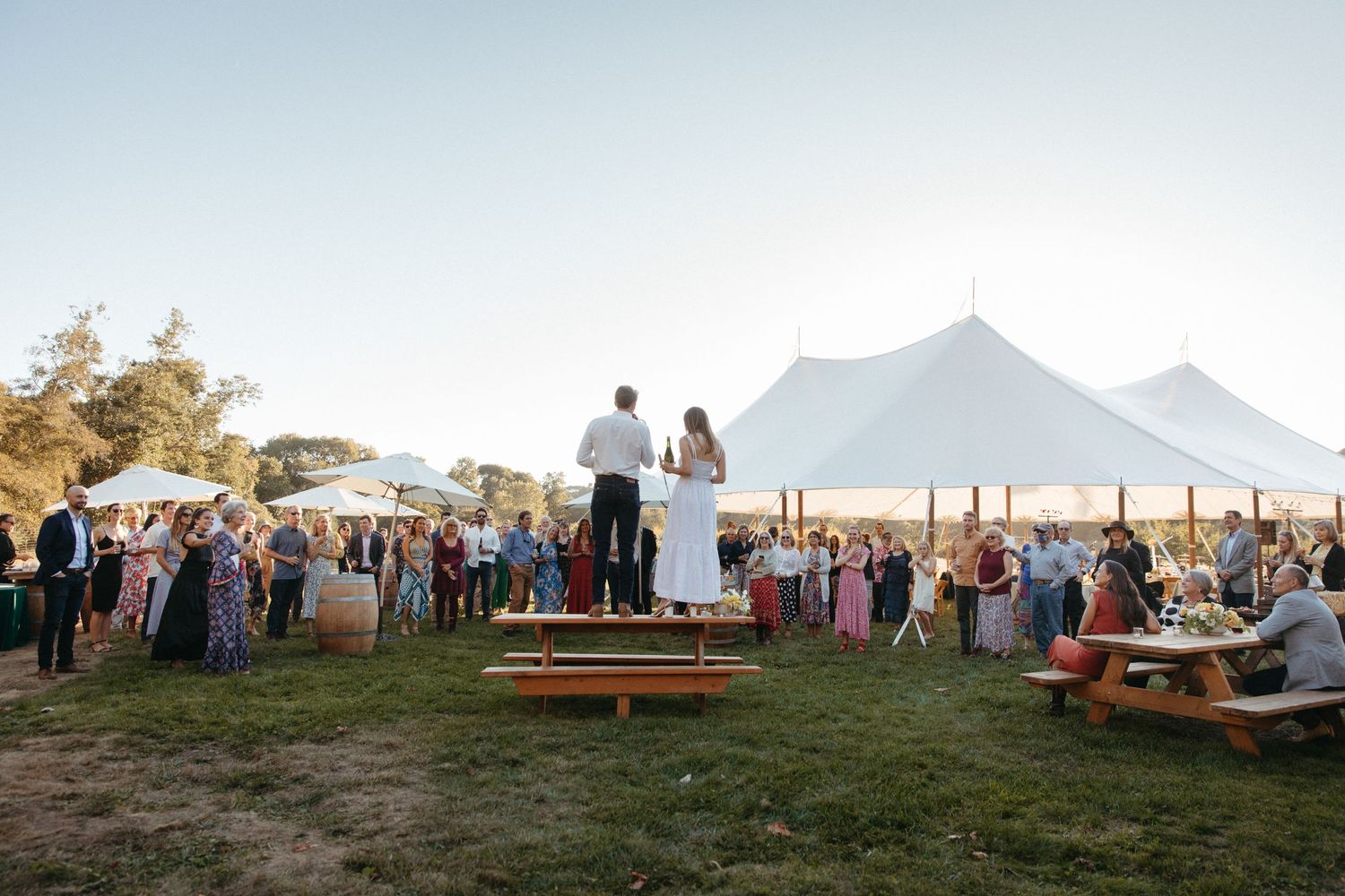 A large outdoor event with guests gathered under a white tent during golden hour in a scenic mountain setting.