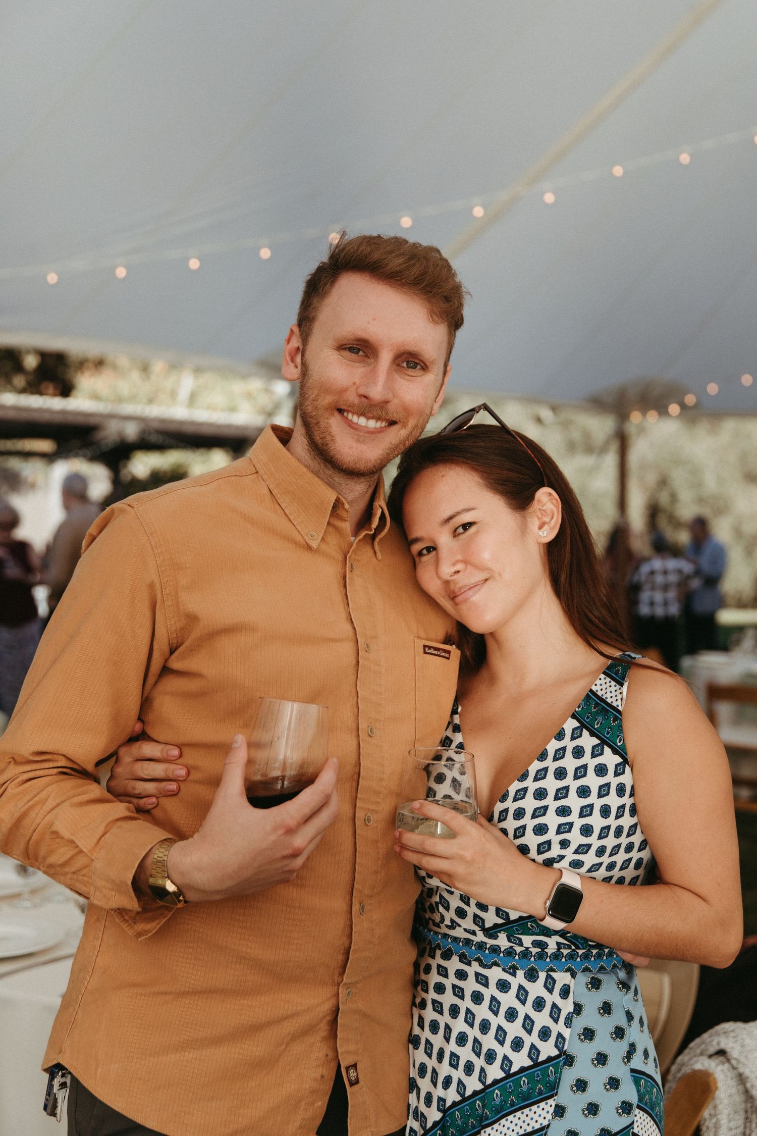A couple enjoys wine at an outdoor event under string lights at dusk.