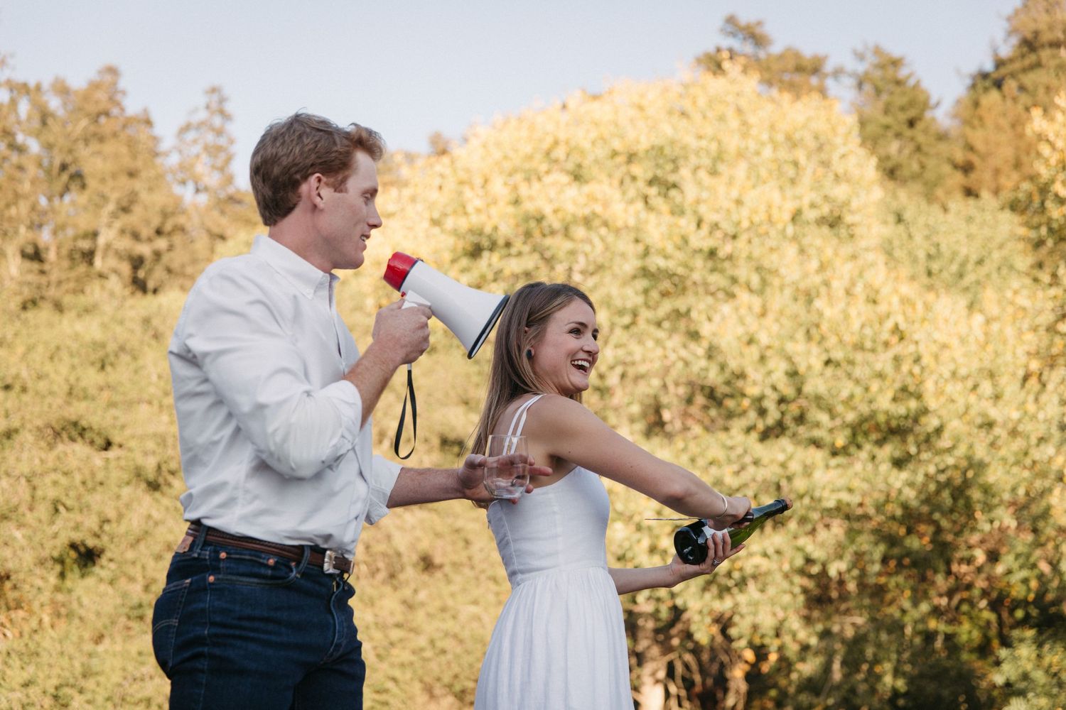 Two people celebrating with megaphones on an outdoor stage with golden fall foliage behind them.