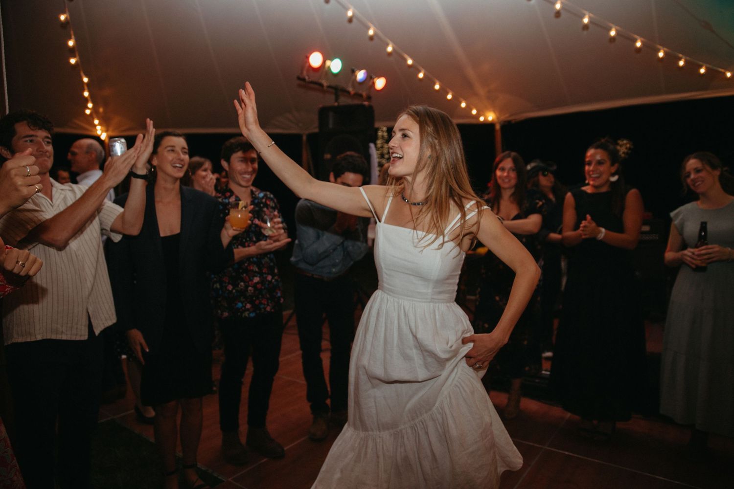 Person in white dress dancing energetically at a reception under string lights.