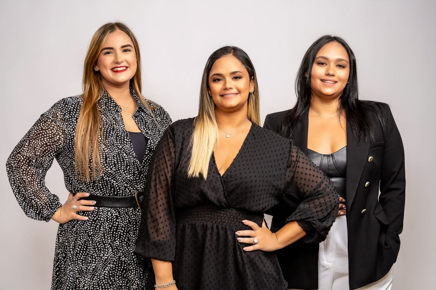 Three women posing together while wearing stylish black dresses against a grey background.