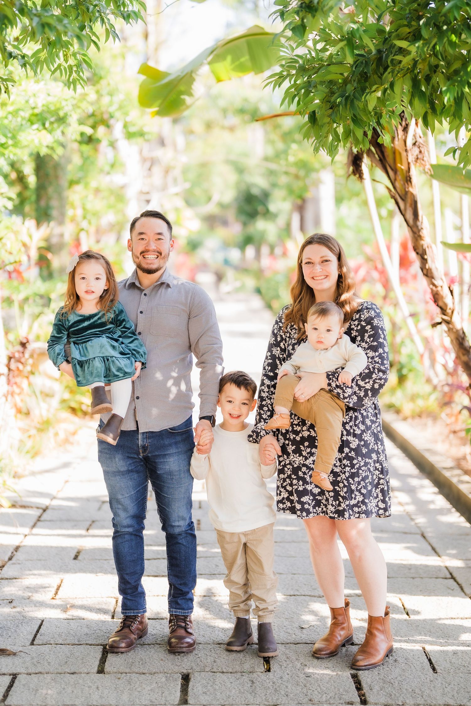 A family of five poses together on a sunlit garden path surrounded by lush greenery.