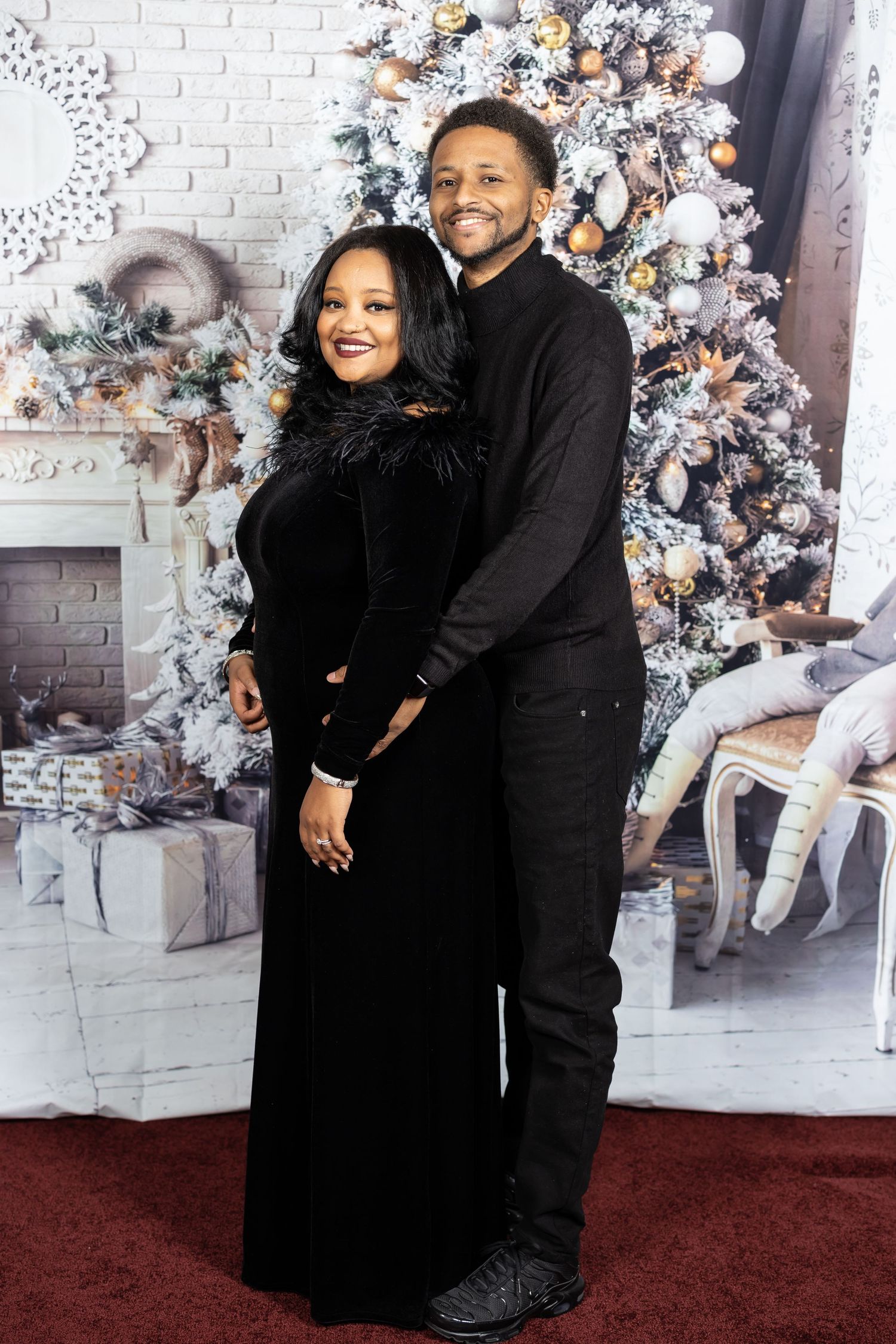 Couple in matching black outfits embraces in front of decorated Christmas scene.