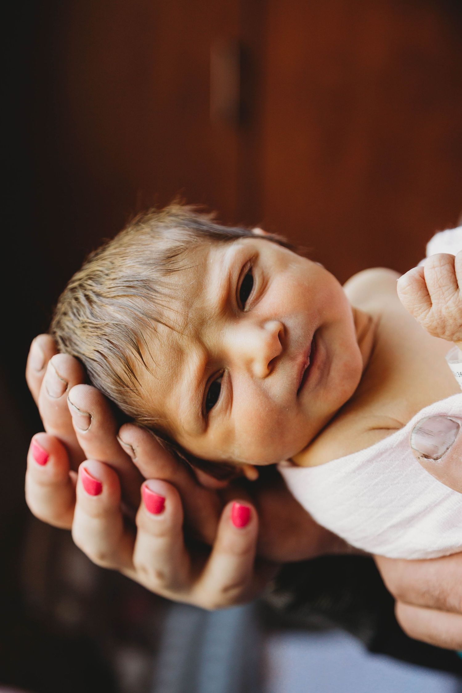 Hands with red painted nails gently cradling a small baby's face.