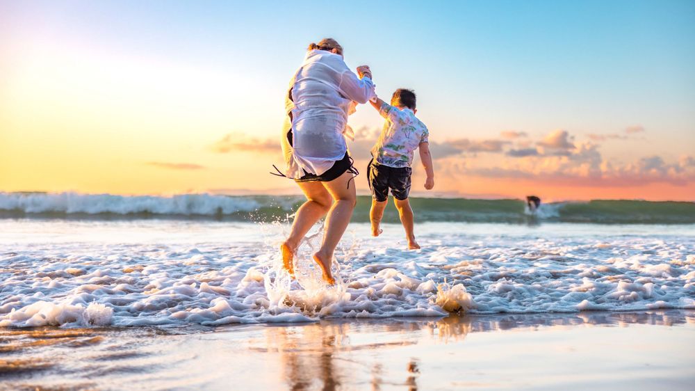 A mother and her son standing at the shoreline of Noosa Beach during sunset jumping over the small waves