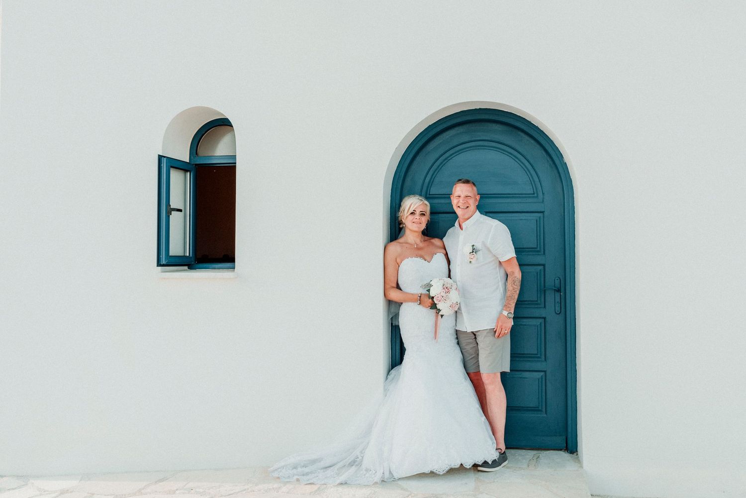 A wedding couple poses in front of a distinctive blue door and white Mediterranean-style architecture.