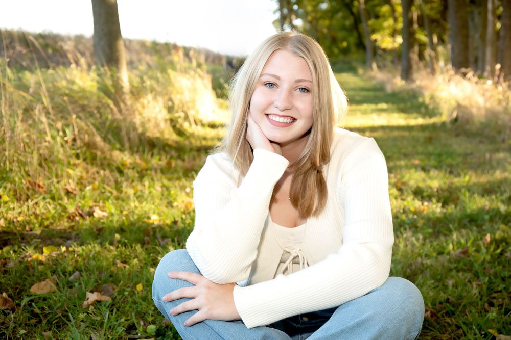 Portrait of someone in white sweater sitting on grass in autumn setting with golden sunlight.