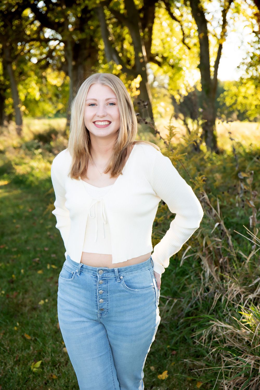 Natural outdoor portrait in white crop sweater and high-waisted jeans with autumn foliage backdrop.