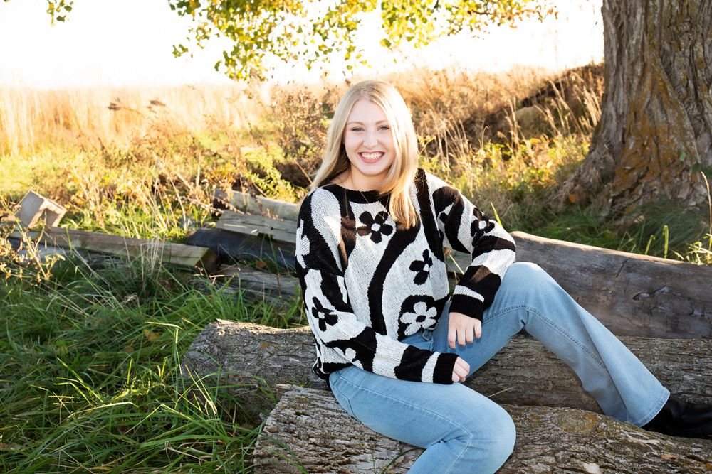 Person in patterned sweater sits on fallen log with autumn foliage in background.