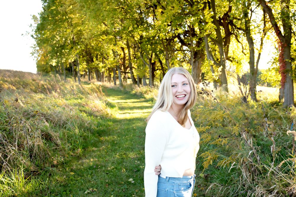 Woman in white sweater poses outdoors on autumn path with golden sunlit trees.