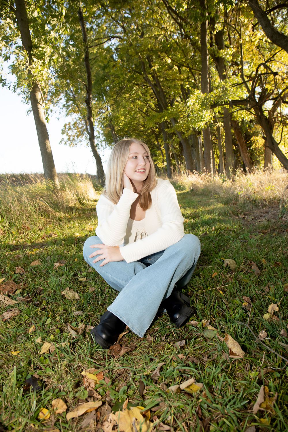 Person wearing white sweater and jeans sits laughing on grass under autumn trees.