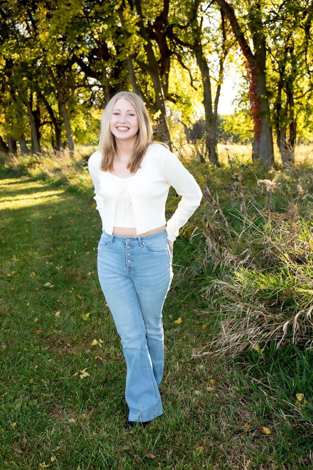 Casual outdoor portrait in white crop top and flared jeans against sunlit trees and grass.