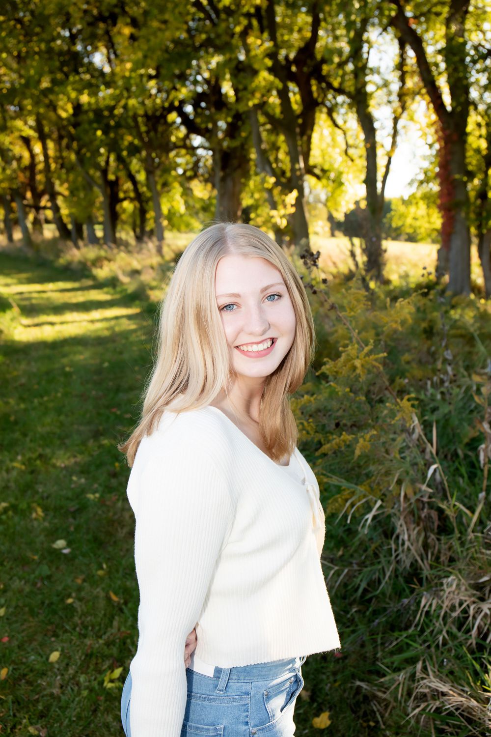 Side-angle portrait in casual white top and jeans with sunny forest background.