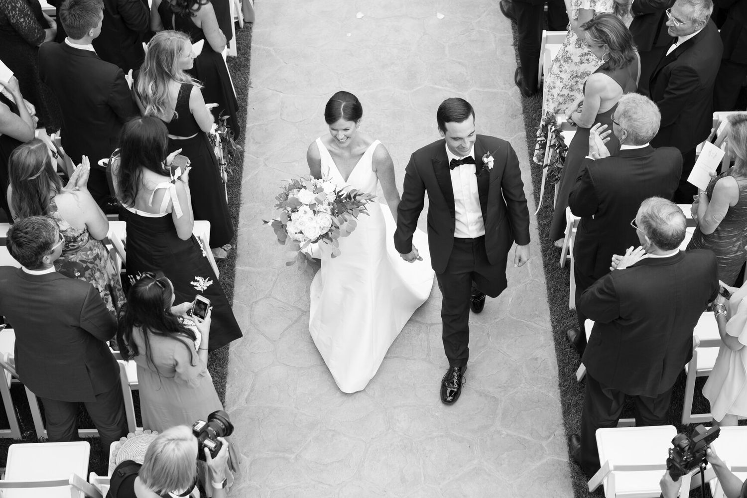 Aerial view of newlyweds walking down the aisle surrounded by guests at an outdoor ceremony.