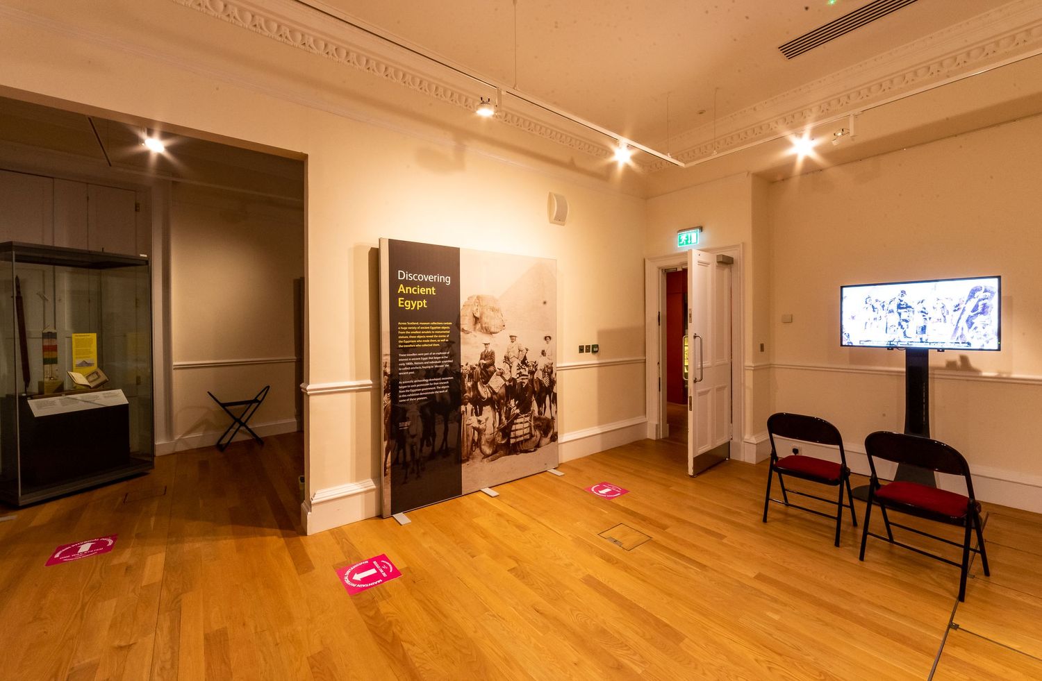 Gallery room with wooden floors, wall displays, and social distancing markers.