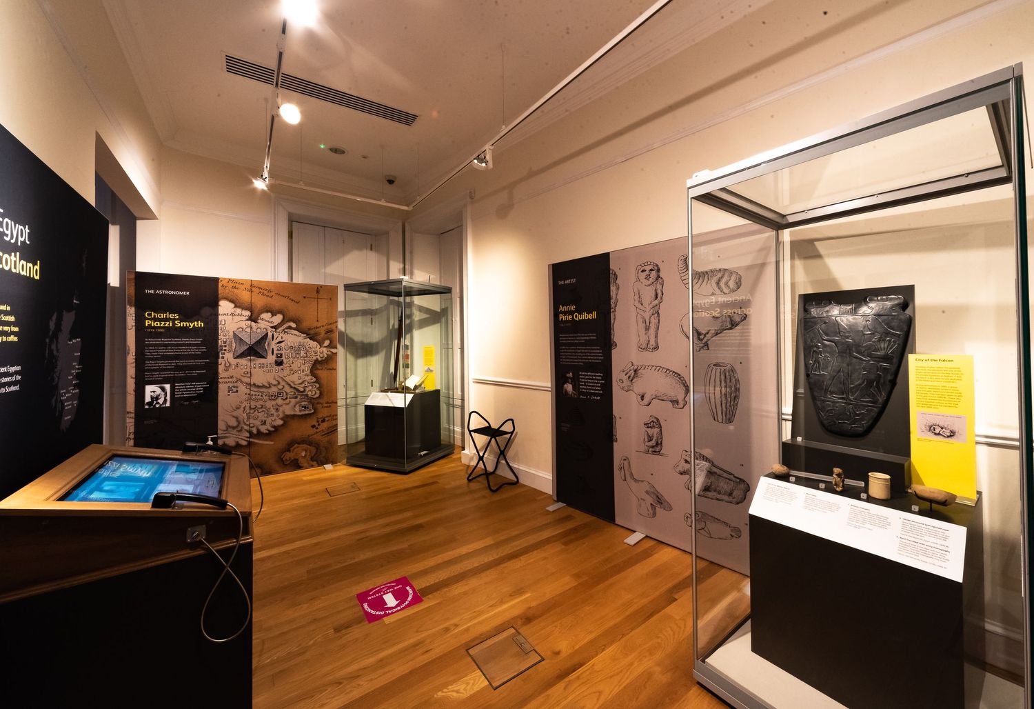 Museum exhibit hall with display cases and historical wall graphics on polished hardwood floor.