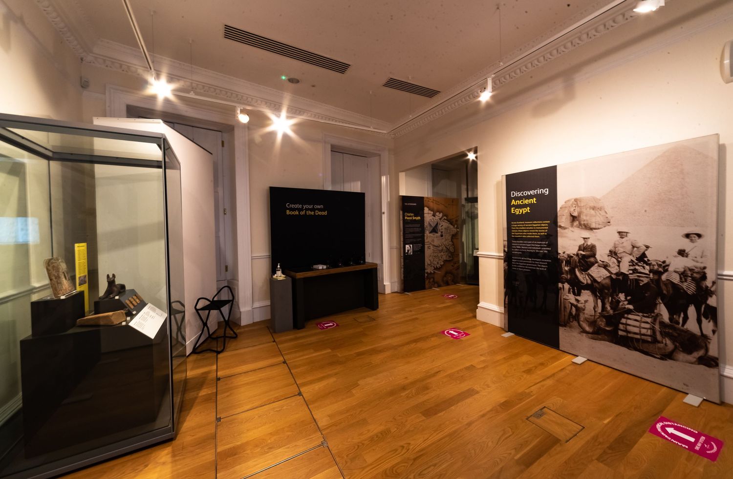 Museum exhibition room with display cases and wall panels on hardwood floors under track lighting.