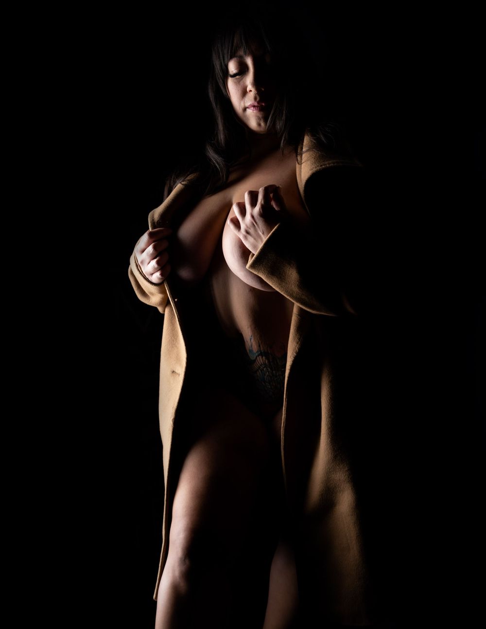 Sensual silhouette in dramatic lighting shows shadowed figure in beige coat against dark background.