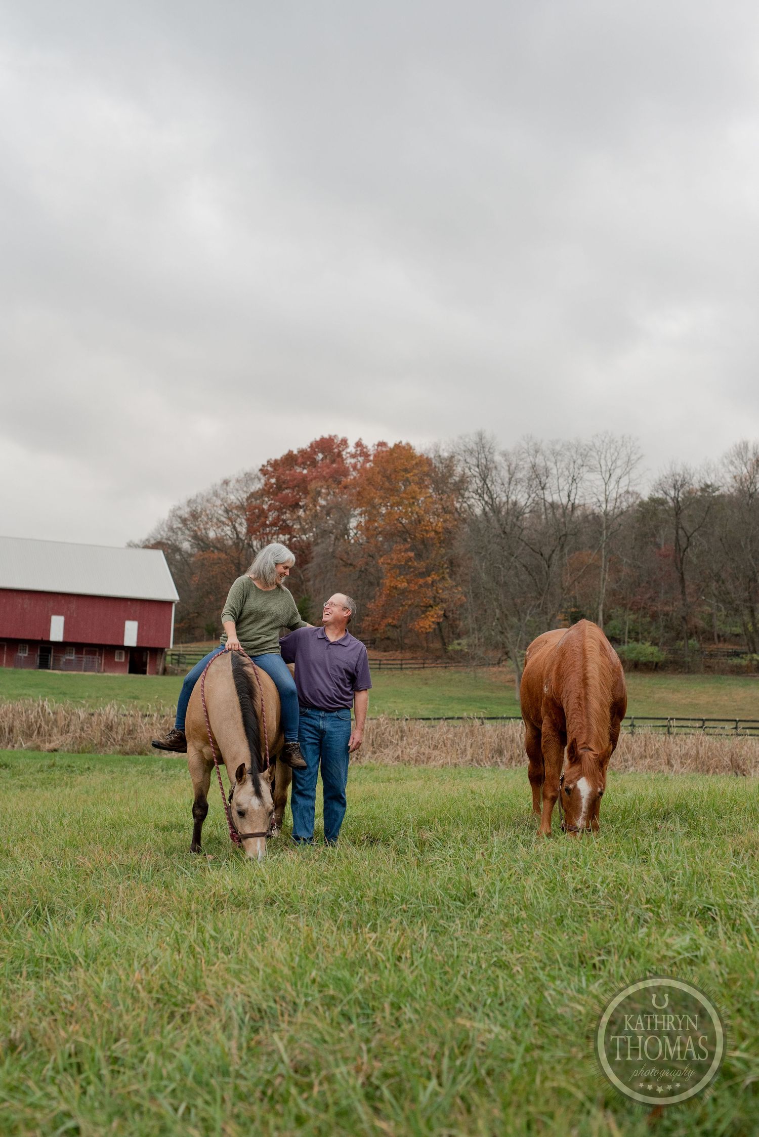 Two people stand with horses grazing in a pastoral setting with red barn and fall colors.