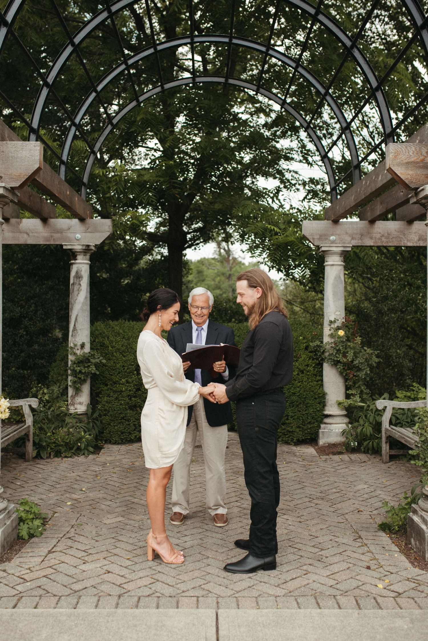 An intimate wedding ceremony takes place under an arched garden structure with stone columns.